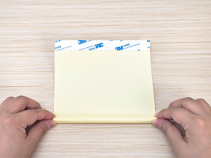 Two hands are shown beginning to roll a piece of pale yellow paper or cardstock on a light woodgrain table. The paper is oriented horizontally, and a strip of 3M double-sided tape is visible along the top edge. The hands are holding the bottom edge of the paper and starting to curl it upwards to form a tube or roll.
