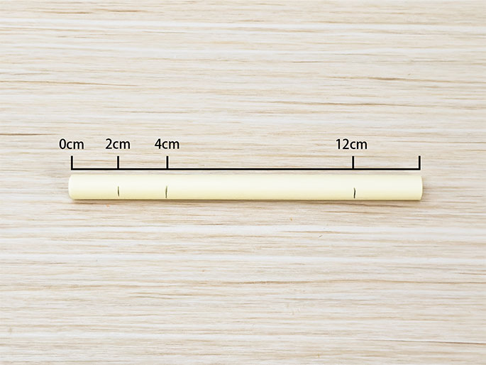 A pale yellow cylindrical object, likely made of paper or cardstock, rests on a light woodgrain surface. It is positioned horizontally and measures 12cm in length, as indicated by a line and label above it. Smaller vertical marks on the line denote 0cm, 2cm, and 4cm intervals. There are faint, small vertical lines drawn on the cylinder itself at approximately the 2cm, 4cm, and 10cm points.