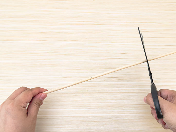 A pair of hands is shown cutting a thin wooden dowel with scissors. The dowel is positioned diagonally across a light woodgrain surface. One hand holds the dowel near the left end, while the other hand holds the scissors, which are in the process of cutting the dowel near the center.