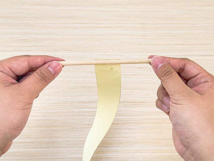 Two hands hold a thin wooden dowel horizontally over a light woodgrain background. Attached to the dowel, and hanging down below it, is a piece of pale yellow paper or cardstock. The paper is slightly curved and appears to be attached to the dowel with tape or adhesive.