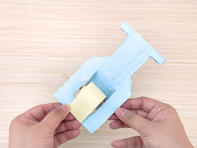 Two hands hold a light blue, roughly hourglass-shaped object made from cardboard or thin wood against a light woodgrain background. A pale yellow roll of paper is positioned within a rectangular opening in the center of the object. A small wooden dowel protrudes from the left side of the object. One hand is gently touching the yellow paper roll. The overall appearance suggests a simple handmade toy or model.
