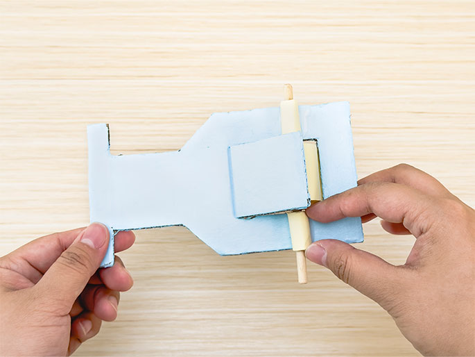 Two hands are manipulating a light blue cardboard or thin wood construction against a light woodgrain background. The construction is roughly hourglass-shaped, and a small wooden dowel is inserted through slots in a rectangular section near the center. The hands are holding the construction and appear to be demonstrating how the dowel can be turned or rotated within the slots.