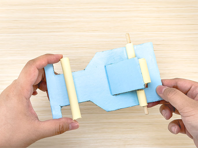 Two hands hold a light blue, roughly hourglass-shaped object made of cardboard or thin wood against a light woodgrain background. A small wooden dowel is inserted through slots on the right side of the central rectangular section. On the left side, a pale yellow cylinder, slightly longer than the dowel, is inserted through corresponding slots. The hands hold the object with the yellow cylinder on the left and the wooden dowel on the right. This likely represents a further stage of construction of a simple handmade toy or model.