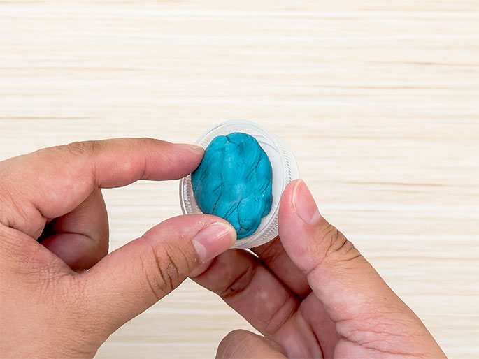 Two hands hold a clear plastic bottle cap containing a small ball of bright teal or turquoise modeling clay or putty. The clay is textured, showing folds and creases. The background is a light woodgrain surface.