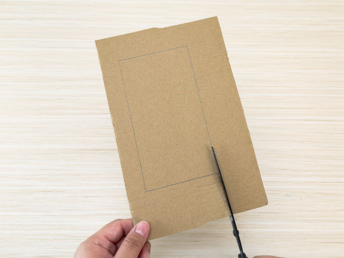 A hand holds a piece of brown cardboard with a rectangular shape drawn on it. A pair of scissors is cutting along the lines of the rectangle. The background is a light woodgrain surface.
