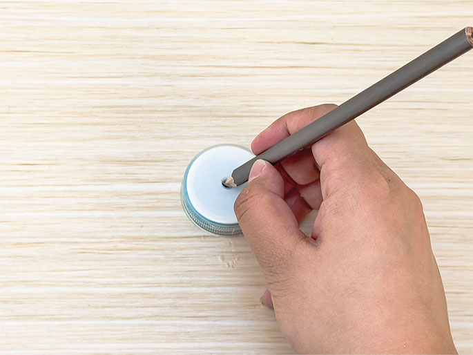 A hand holding a gray pencil is poking the pencil point through a hole in a clear plastic bottle cap. The cap is placed on a light woodgrain surface. The inside of the cap appears to have a light blue or white surface.