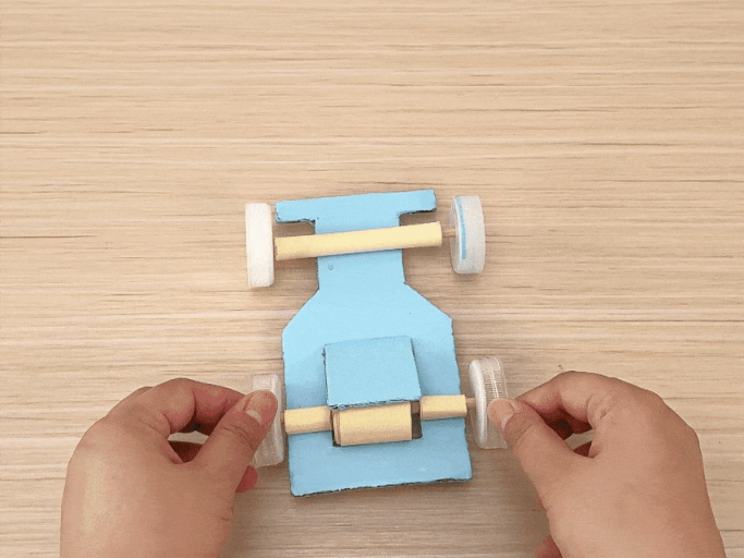 Two hands assemble a small, light blue toy car on a light wooden table. The car's body is made of cardboard, and its axles are constructed from rolled-up paper or thin cardboard tubes. Plastic bottle caps serve as the wheels. The hands attach the bottle cap wheels to the axles, completing the car's construction. The video focuses on the process of attaching the wheels, showcasing the simple materials and design.