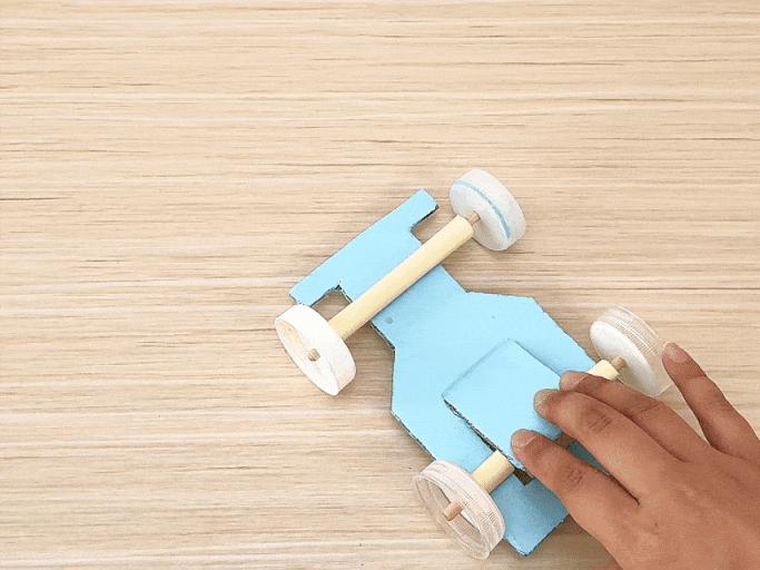 A small, light blue toy car, crafted from cardboard and plastic bottle caps for wheels, rests on a light wooden table. The car features exposed axles made from what appears to be rolled-up paper or thin cardboard tubes. A hand briefly enters the frame at the bottom right. The car does not move, and a popping sound is heard, possibly indicating an off-screen action or the sound of something being placed on the table.