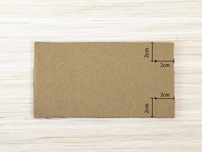A rectangular piece of brown cardboard lies on a light woodgrain surface. Three points are marked along one of the shorter edges, each 2cm apart, as indicated by lines and labels. This suggests markings for folding or cutting.