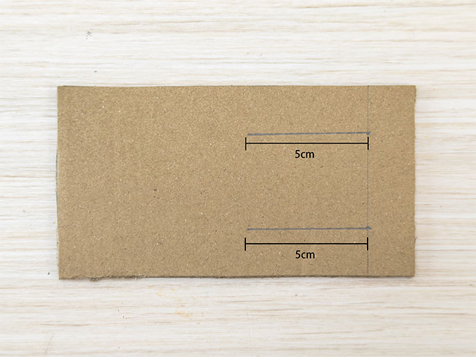 A rectangular piece of brown cardboard is placed on a light woodgrain surface. Two parallel lines, each 5cm long as indicated by labels, are lightly drawn on the cardboard. These markings suggest planned cuts or folds.