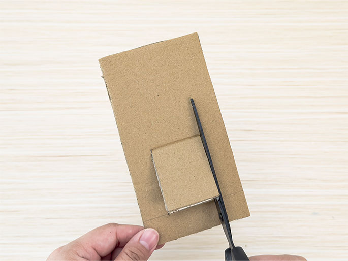 A hand holds a piece of brown cardboard on a light woodgrain surface. A small rectangular flap has been cut on three sides and folded outward. Scissors are cutting along the final uncut line of the flap.
