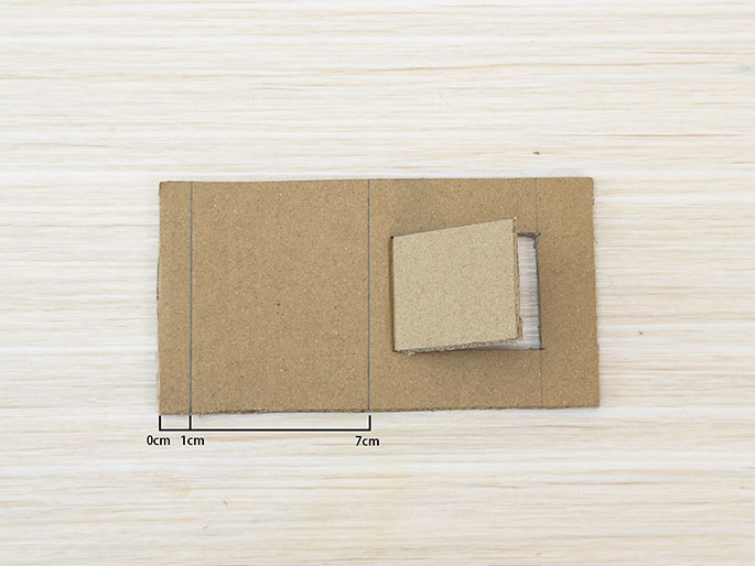 A piece of brown cardboard rests on a light woodgrain table. A small rectangular flap is partially cut out and folded open. A vertical line is lightly drawn on the cardboard to the left of the flap. A horizontal line at the bottom edge shows a measurement of 7cm, with 0cm and 1cm markings to the left, indicating the position of the vertical line. This suggests further cutting or folding along the marked lines.
