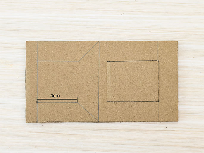 A rectangular piece of brown cardboard is on a light woodgrain surface. Lines are drawn on it, outlining a smaller rectangle on the right side and a larger, irregular shape on the left. A line labeled 4cm indicates the width of the bottom section of the larger shape. These markings appear to be guides for cutting or folding.
