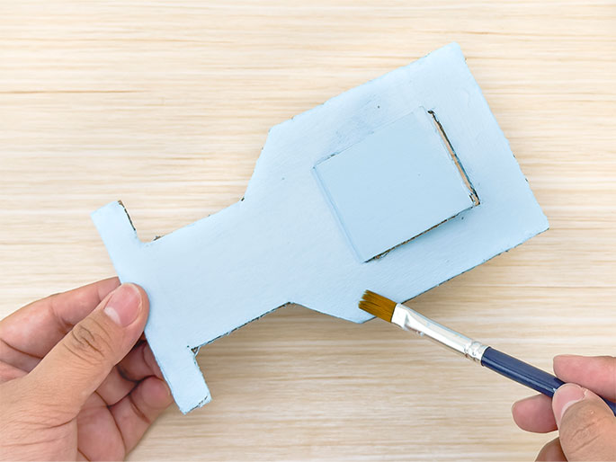 Two hands are working on a light blue painted cardboard cutout on a light woodgrain surface. The cutout, resembling a simplified camera or projector, is being painted with a small brush held in the right hand. The left hand holds the cutout steady. The brush is applying paint to the lower right section of the main body of the object.