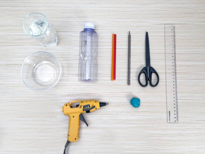 An assortment of craft supplies is laid out on a light wood grain table. The items include a glass of water, an empty clear plastic bottle, two colorful straws or sticks (one orange, one red), a pencil, a pair of scissors, a clear plastic ruler, a yellow hot glue gun, and a small lump of teal or blue-green modeling clay or putty. These materials appear ready for use in a DIY or crafting project.