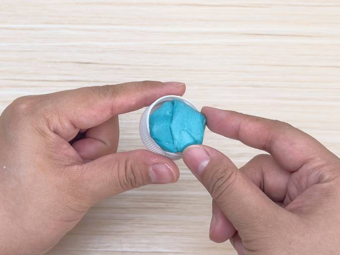 Two hands hold a white plastic bottle cap filled with a turquoise or teal-colored putty or modeling clay. The hands are positioned against a light wood grain background. They appear to be pressing or shaping the putty within the cap.
