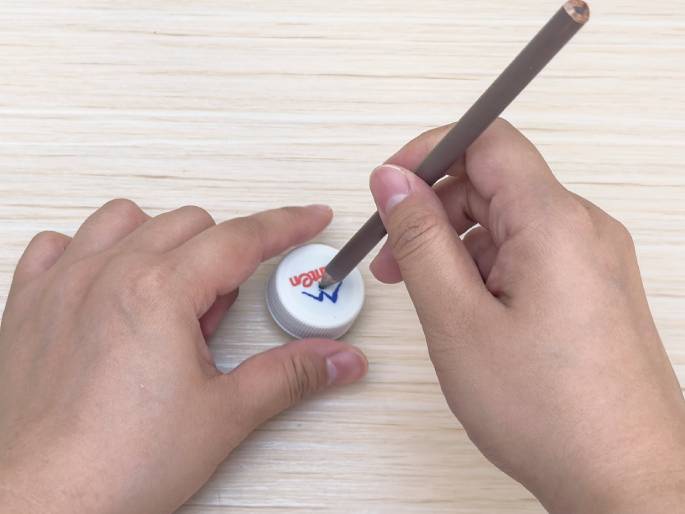 Two hands manipulate a white plastic bottle cap and a pencil on a light wood grain surface. One hand holds the cap steady while the other uses the pencil to mark or draw on the top of the cap. The cap has some pre-existing text or a logo visible.