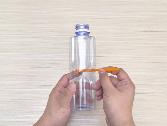 Two hands hold a clear, empty plastic bottle and a small orange stick against a light wood grain background. One hand holds the bottle while the other positions the orange stick horizontally across the middle section of the bottle. This suggests measuring or marking for a craft or DIY project.