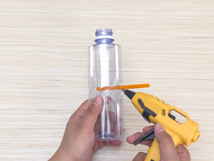 Two hands work with a clear, empty plastic bottle and a yellow hot glue gun on a light wood grain background. An orange stick is attached horizontally across the middle of the bottle. One hand holds the bottle while the other applies hot glue from the glue gun to secure the stick. This image depicts a step in a crafting or DIY project.