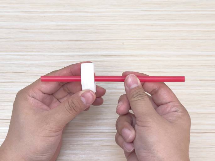 Two hands hold a red plastic drinking straw and a white plastic bottle cap on a light wood grain background. The straw is inserted through the bottle cap, creating an axle-like assembly. This suggests a stage in constructing a simple toy or craft project.