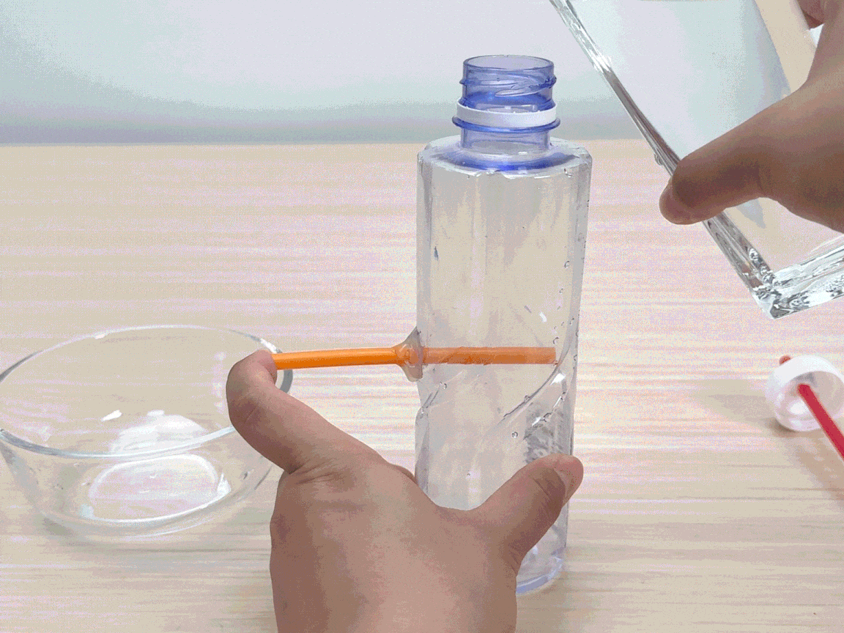 A clear plastic bottle, partially filled with water, has a small orange stick protruding from its side. A hand pours more water into the bottle from a clear glass container. A clear glass bowl sits on the light wooden table surface near the bottle. In the background, a red straw is inserted through a white plastic bottle cap. This scene appears to demonstrate a science experiment or DIY project, possibly exploring concepts like water flow or pressure.