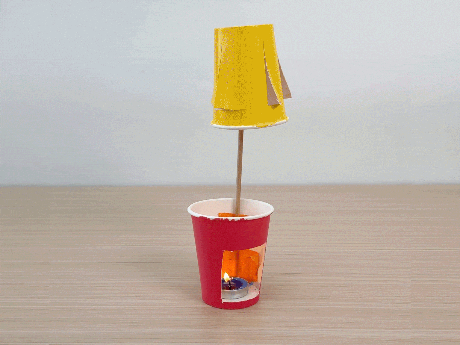 A small, homemade hot air balloon spins above a lit tea candle. The balloon is made from a yellow paper cup with cutouts, attached to a stick inserted into a red paper cup. The tea candle sits inside the red cup, providing the heat source. This demonstrates a simple science experiment.