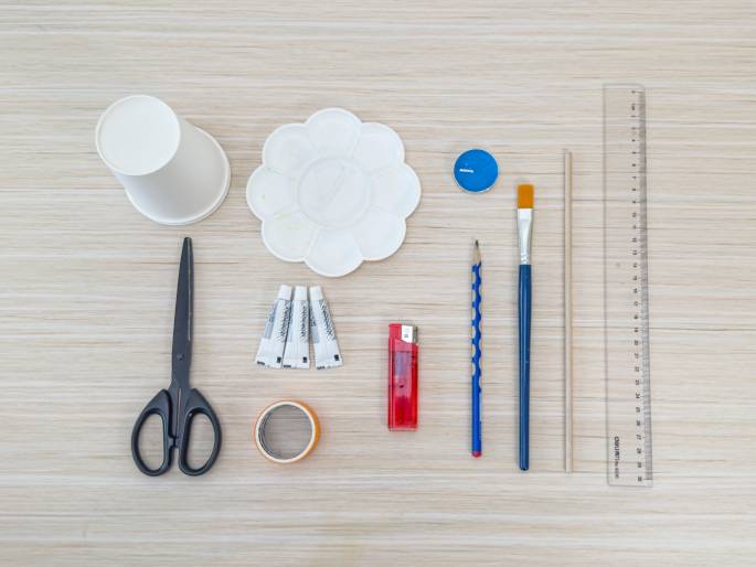 A variety of craft and stationery items are laid out on a light wood-grain table. These include an upside-down white paper cup, a white flower-shaped paint palette, a small container of blue paint, a paintbrush with a blue handle and yellow bristles, a pencil with a blue textured grip, a long thin wooden dowel, a clear plastic ruler, a red lighter, three small tubes of white glue, a pair of black scissors, and a roll of orange masking tape. The arrangement suggests preparation for a craft project or DIY activity.