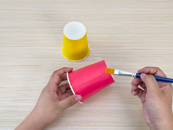 On a light-colored wooden surface, two hands are engaged in a painting activity. A left hand holds a small pink paper cup, tilted slightly, while a right hand applies orange paint to the cup's exterior using a paintbrush with a blue handle and yellow bristles. Another yellow paper cup sits upright in the background. The scene suggests a simple arts and crafts project.