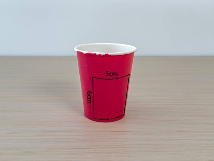 A bright pink paper cup sits on a light wood-grain table. A rectangular area is marked on the cup with black lines and labeled with dimensions: 5cm across the top and 6cm down the side. This image appears to be providing measurements for a craft or DIY project.