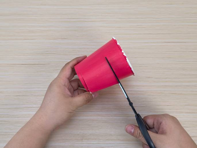 Two hands are cutting a bright pink paper cup on a light wood-grain table. The left hand holds the cup at an angle, while the right hand uses black scissors to make a vertical cut down the side. This image likely depicts a step in a craft or DIY project.