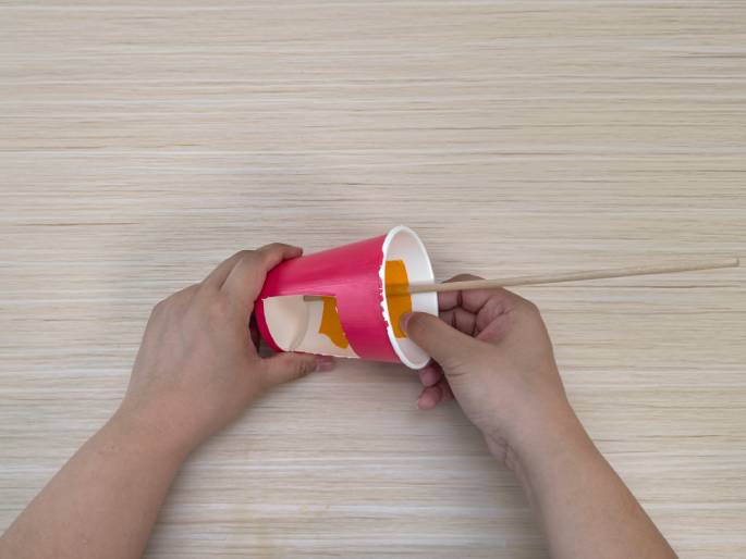 Two hands are working with a modified pink paper cup on a light wood-grain table. A rectangular hole has been cut into the side of the cup, and a thin wooden dowel is being inserted through this opening. Orange material is visible inside the cup around the hole. This image appears to be a step in constructing a craft or DIY project.