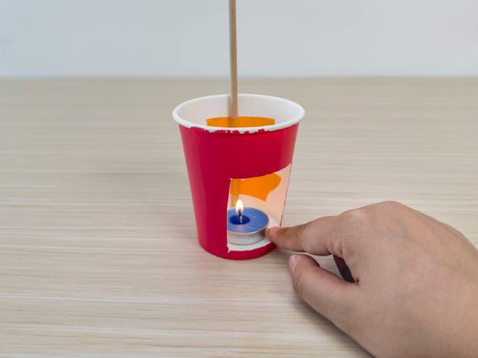 A small, lit tea light candle sits inside a modified pink paper cup on a light wood-grain table. A rectangular opening has been cut into the cup's side to reveal the candle's flame. A thin wooden dowel extends upwards from the cup's opening. A hand gestures towards the candle, with a finger pointing near the flame. Orange material is visible inside the cup, behind the candle. This image appears to showcase a completed DIY or craft project, possibly a small lantern.