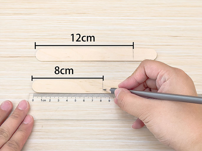 Two wooden craft sticks lie on a light wood surface. A hand uses a pencil and a clear ruler to mark a point on the lower stick at 8cm. A line above the first craft stick indicates a length of 12cm. The scene suggests the beginning steps of a crafting project involving measurement.