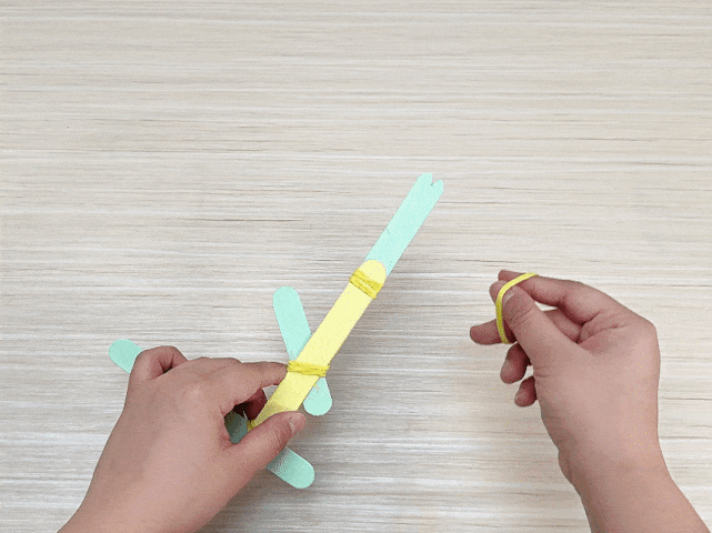 On a light wood surface, two hands are assembling a craft stick catapult. One hand holds the catapult, which consists of a yellow stick as the base, a light green support stick perpendicular at the bottom, and another light green, notched stick on top. These are secured with yellow rubber bands. A small piece of light green craft stick is wedged between the top and base sticks. The other hand holds a separate yellow rubber band, ready to complete the catapult assembly.