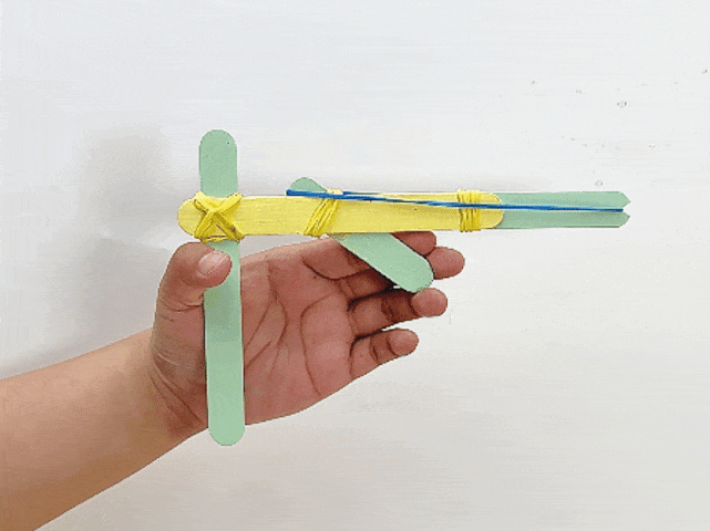 A hand holds a completed craft stick catapult against a plain white background. The catapult is constructed with a yellow stick as the base and light green sticks forming the support and launching arm. Yellow rubber bands secure the structure at the intersection points. A small green stick piece is wedged between the launching arm and the base. A thin blue rubber band is stretched along the top of the catapult, ready to launch a projectile.