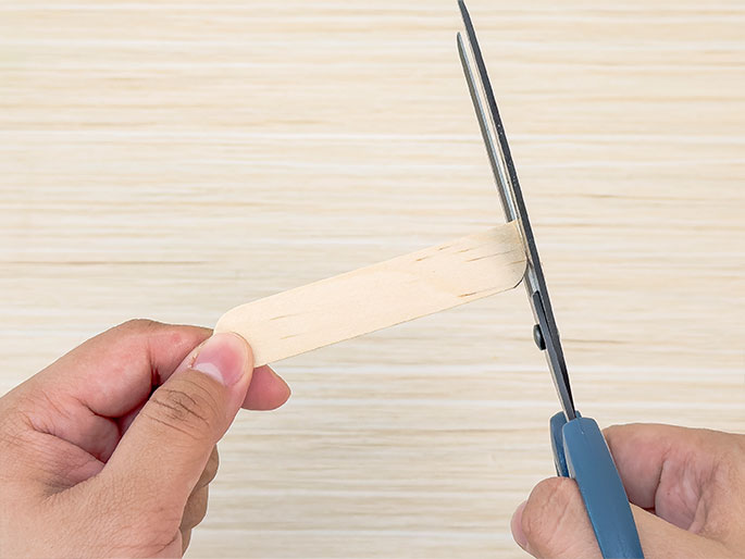Two hands are shown against a light wood background. One hand holds a wooden craft stick, while the other hand uses scissors to cut the end of the stick. The image suggests a DIY or crafting activity.
