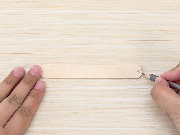 A wooden craft stick rests on a light wood surface. One hand holds the stick while another hand uses a pencil to draw a small notch on the right end. The scene suggests a crafting activity, possibly marking the wood for cutting or shaping.
