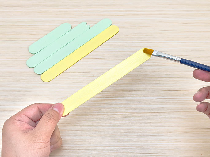On a light wooden surface, several craft sticks are visible. Some are painted a light green, while others are yellow. A hand holds a yellow craft stick as another hand uses a paintbrush to apply yellow paint to it. The image captures the process of painting craft sticks for a craft project.