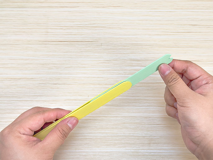 Two hands hold two painted craft sticks – one yellow and one light green – against a light wood background. The green stick is placed on top of the yellow stick, and they are being held together. The green stick has a notched end. The image suggests a stage in a crafting process where the sticks are being combined.