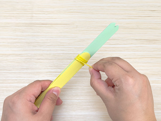 Against a light wood background, two hands manipulate a pair of craft sticks. A yellow rubber band is being wrapped around the area where a light green and a yellow craft stick overlap. The green stick has a notched end. This image depicts a step in a crafting process, likely creating a small catapult or similar device.
