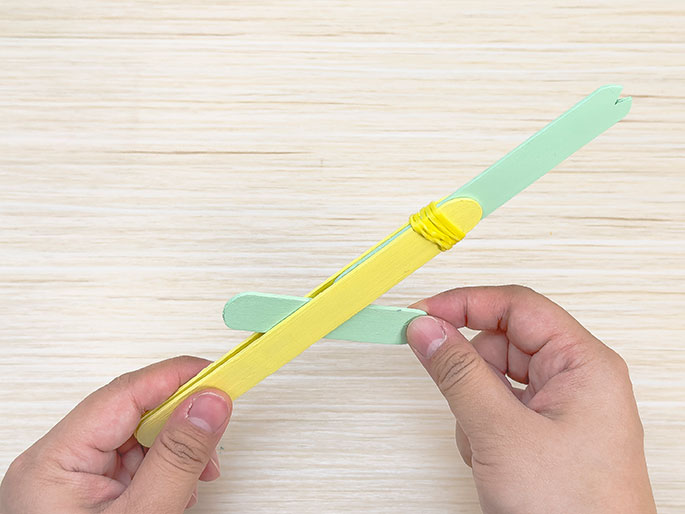 Two hands are assembling a small catapult made of craft sticks on a light wood background. A yellow craft stick and a light green one are held together with a yellow rubber band at one end. A second light green stick is wedged between the first two. The green stick on top has a notched end. The image shows a stage in constructing a craft stick catapult.