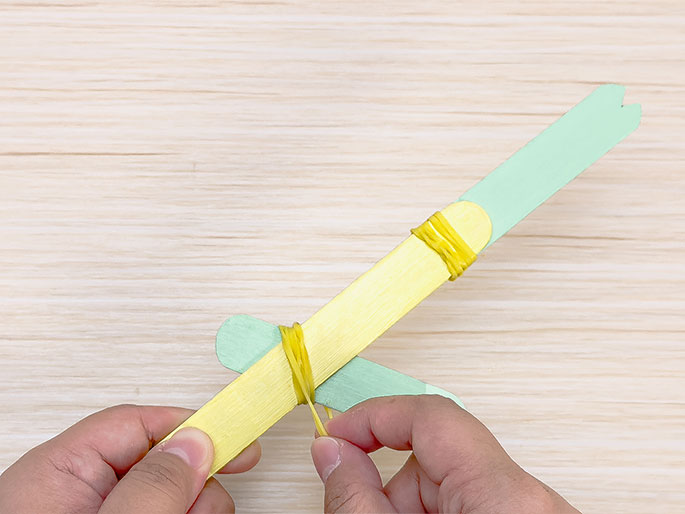 Against a light wooden backdrop, two hands are shown constructing a craft stick catapult. A yellow craft stick is positioned diagonally, with a light green stick wedged perpendicularly across it. Yellow rubber bands are wrapped around both intersection points of the sticks. The green stick attached to the top end of the yellow stick has a notched end. The image illustrates a step in the process of making a simple catapult using craft sticks and rubber bands.