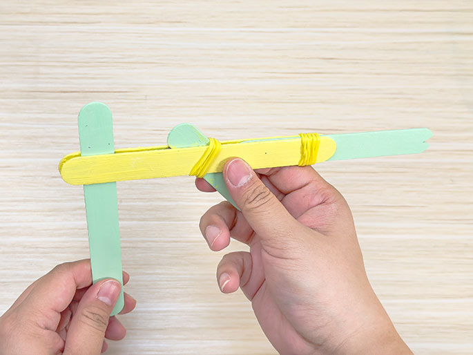 Against a light wood background, two hands hold a nearly completed craft stick catapult. A yellow stick forms the base, with a light green stick perpendicular at the bottom and another light green stick on top, secured with yellow rubber bands. The top green stick has one end notched and extends past the yellow stick, forming the projectile launching arm. The other end of the top green stick is wedged under the yellow stick, creating the tension mechanism. A smaller piece of a green craft stick is wedged between the yellow stick and the launching arm. The image showcases the final stages of assembling the catapult.
