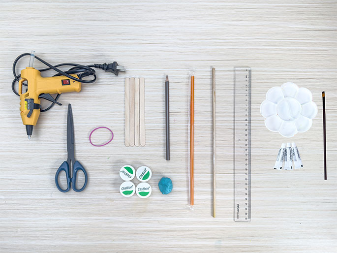 An assortment of craft supplies lies arranged on a light woodgrain work surface. These include a yellow hot glue gun with its power cord, a pair of scissors, a pink rubber band, three wooden craft sticks, a gray pencil, an orange straw or plastic rod, a thin wooden dowel, a clear plastic ruler, a white flower-shaped paint palette, a small paintbrush, three small tubes of glue, four plastic bottle caps, and a small lump of teal or turquoise modeling clay. The items appear to be laid out in preparation for a craft project.