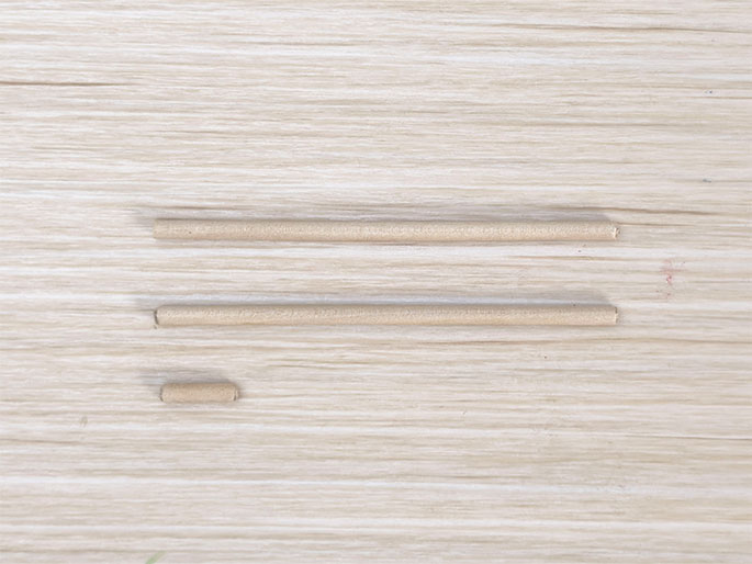 Two thin, light-colored wooden dowels of equal length are positioned horizontally on a light woodgrain surface. A shorter segment of a similar dowel is placed below them, centered. This image likely depicts materials prepared for a craft or construction project.