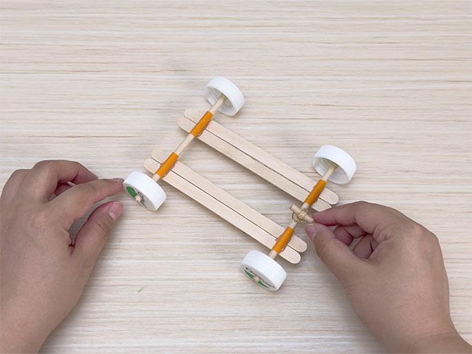 Two hands are shown manipulating a small toy car constructed from craft materials on a light woodgrain surface. The car's frame is made from wooden craft sticks, the wheels are white plastic bottle caps, and orange tape or bands are wrapped around the axles. Thin wooden dowels serve as axles, connecting the wheels to the frame. The hands appear to be in the process of attaching or adjusting a small piece of the car's undercarriage.