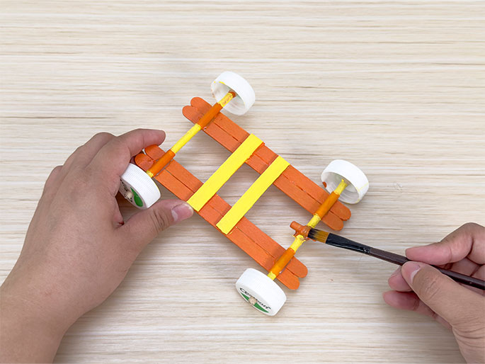 Two hands are working on a toy car made of craft materials on a light woodgrain table. The car's body is constructed from orange-painted wooden craft sticks, and it has four wheels made from white plastic bottle caps. Yellow stripes decorate the top of the car's frame. One hand holds the car while the other hand uses a paintbrush to apply orange paint to a detail on the back of the vehicle. This image captures the final stages of painting the toy car.
