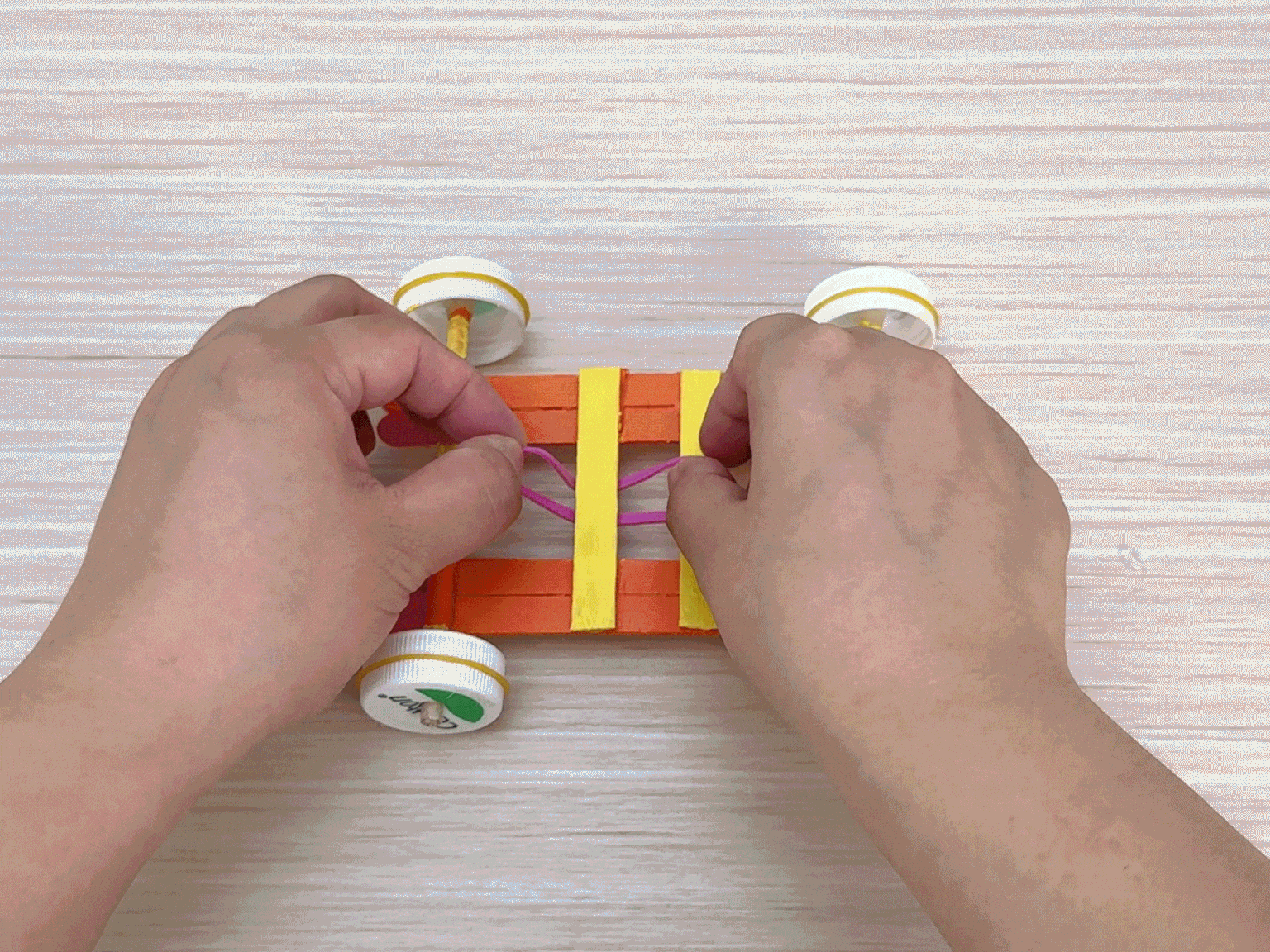Two hands work on a small toy car made of orange and yellow popsicle sticks on a light wooden surface. The car has four wheels, each made from a white plastic bottle cap attached to a small wooden axle. The hands stretch a pink rubber band across the center of the car's frame, likely creating the mechanism for propelling the vehicle. The video focuses on the process of attaching the rubber band motor.