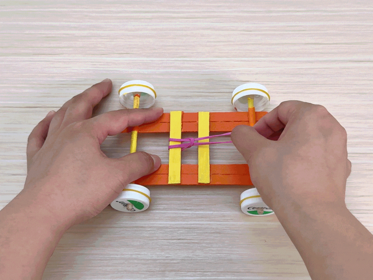 Two hands manipulate a small toy car constructed from orange and yellow popsicle sticks on a light wooden table. The car has four wheels made from white plastic bottle caps affixed to thin wooden axles. A pink rubber band is wrapped around the center of the car's frame, between the axles, likely serving as the car's motor mechanism. The hands adjust and tighten the rubber band, preparing the car for propulsion. The video focuses on the final steps of assembling the rubber band motor.