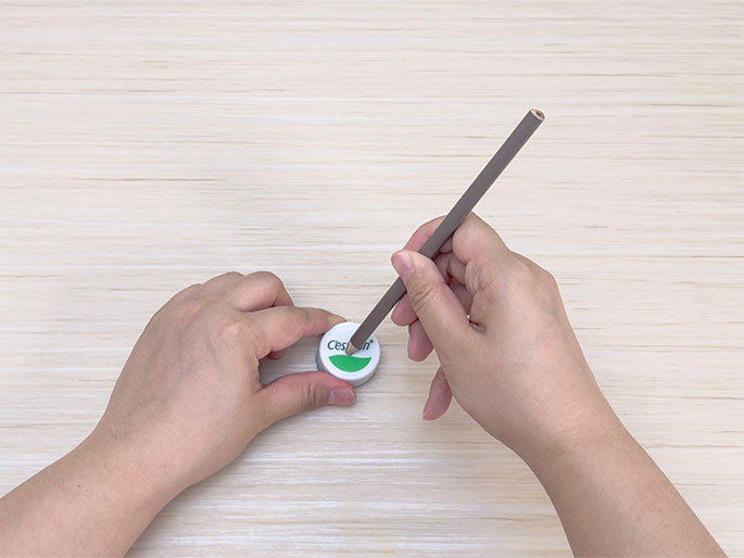 Two hands are shown on a light woodgrain surface. One hand holds a white plastic bottle cap with green lettering. The other hand holds a gray pencil and is using it to mark or draw on the top of the bottle cap.